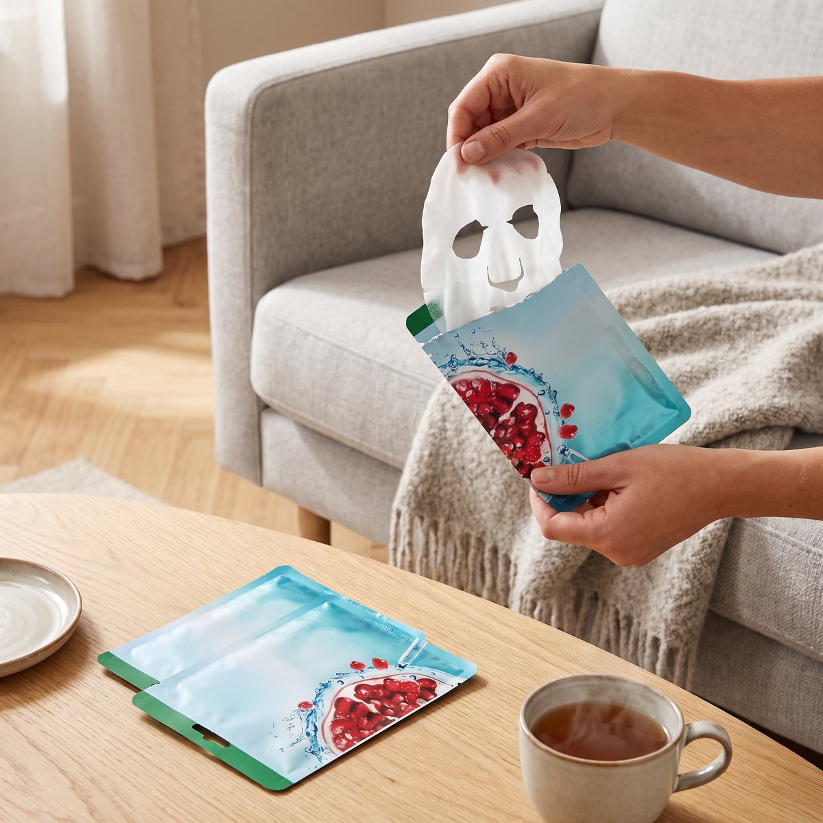 Händer tar en blöt arkmask ur Garnier-påsen i soffan i ett svenskt vardagsrum, med två oöppnade påsar och en kopp te på soffbordet
