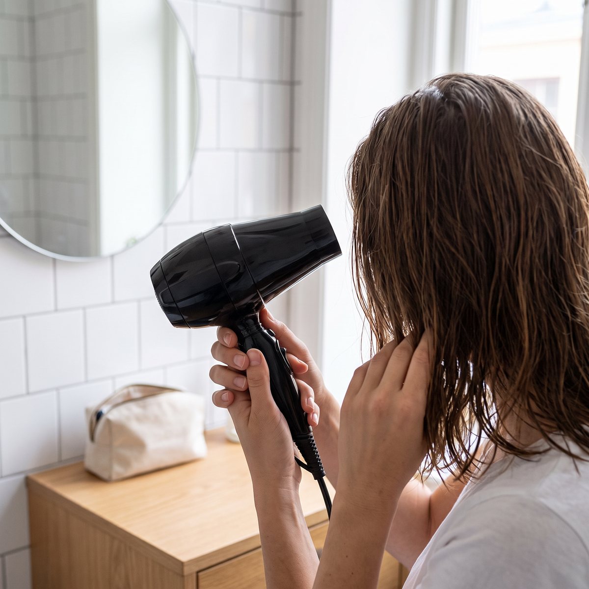 BaByliss PRO Bambino Travel, en liten svart resefön, används för att torka axellångt hår i ett ljust hotellbadrum med vitt kakel