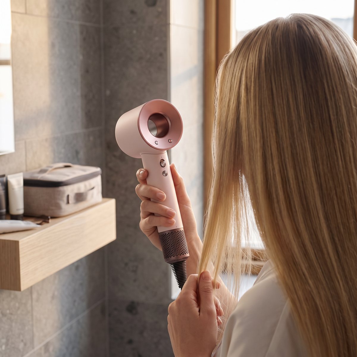 Dyson Supersonic Travel i rosa och koppar torkar långt blont hår i ett hotellbadrum med grå stenkakel