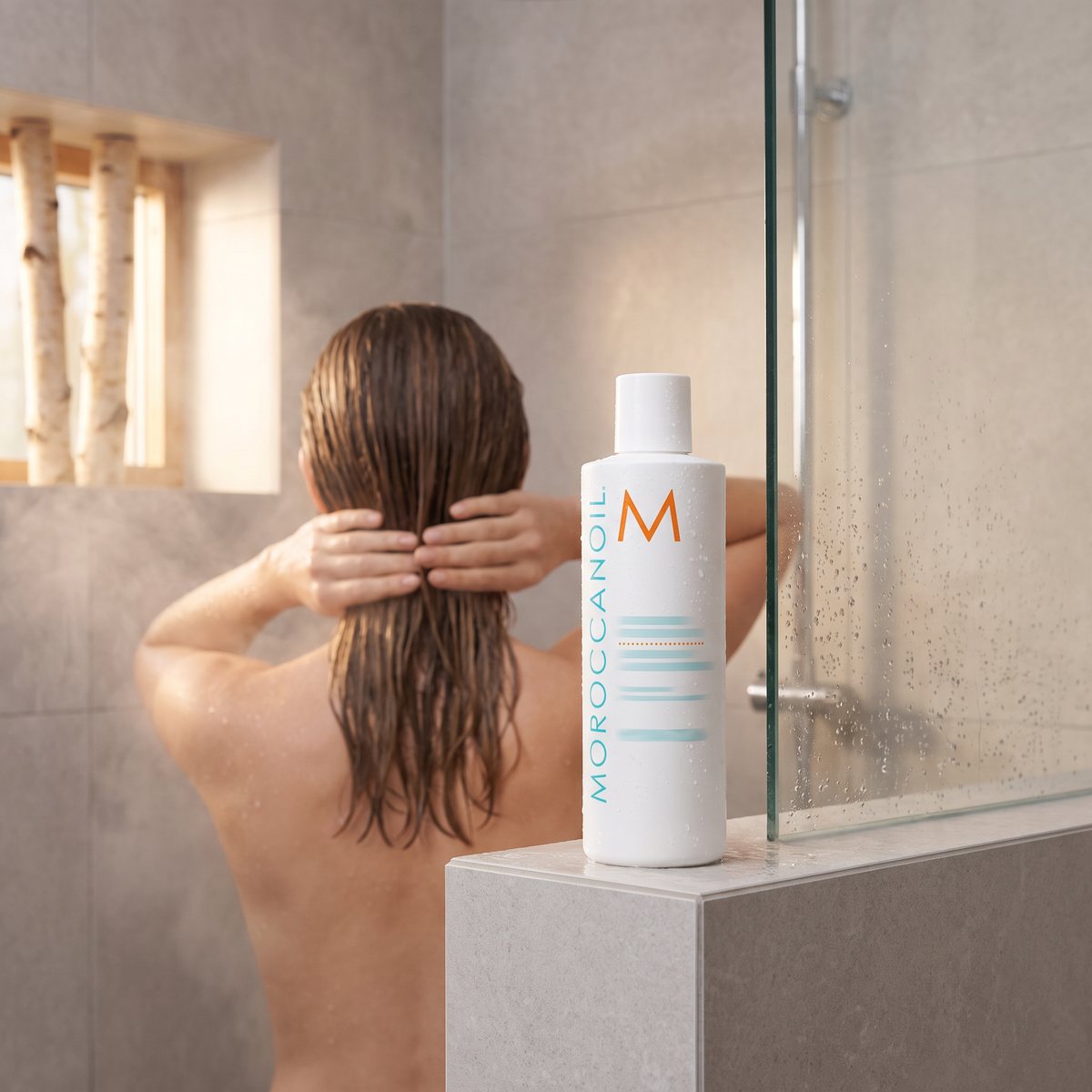 Moroccanoil Hydrating Conditioner på kanten av en duschvägg i ett grått klinkerbadrum, i bakgrunden arbetas balsam in i blött brunt hår, björkar utanför fönstret.