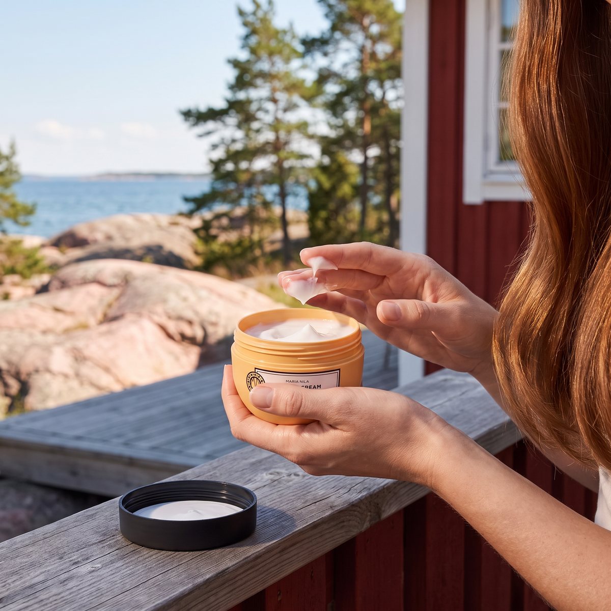 Fingrar tar kräm ur en öppen burk Maria Nila Styling Cream på trädäcket vid en röd stuga i skärgården en solig sommardag