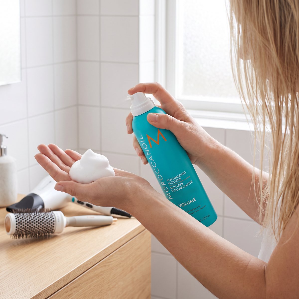 Moroccanoil Volumizing Mousse i handen vid ett handfat i ett ljust svenskt badrum, med en golfbollsstor klick skum i handflatan och rundborste och hårfön på kommoden
