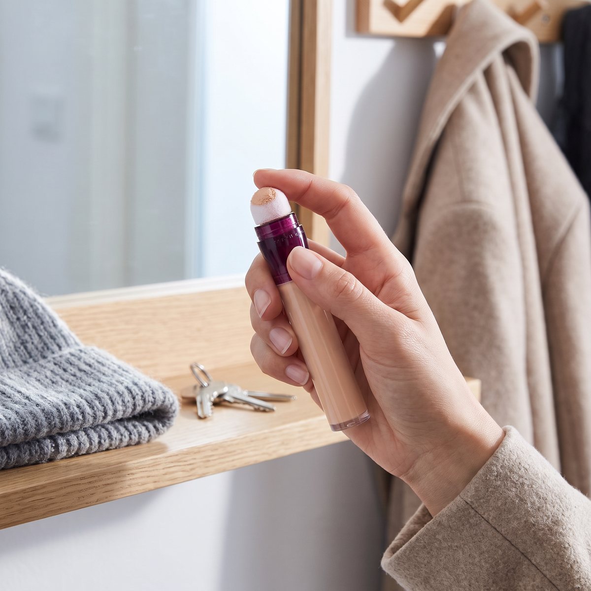 Hand håller Maybelline Instant Anti-Age Eraser med svampspetsen uppåt framför hallspegeln i en svensk lägenhet, med nycklar och mössa på hyllan