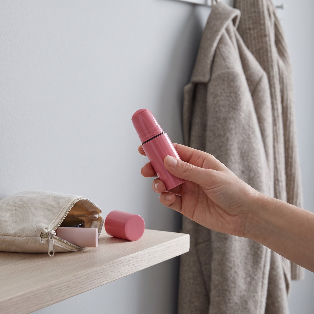 En hand håller ett uppvridet rosa IsaDora Blush Stick vid en hatthylla i en ljus hall, med locket bredvid en necessär.