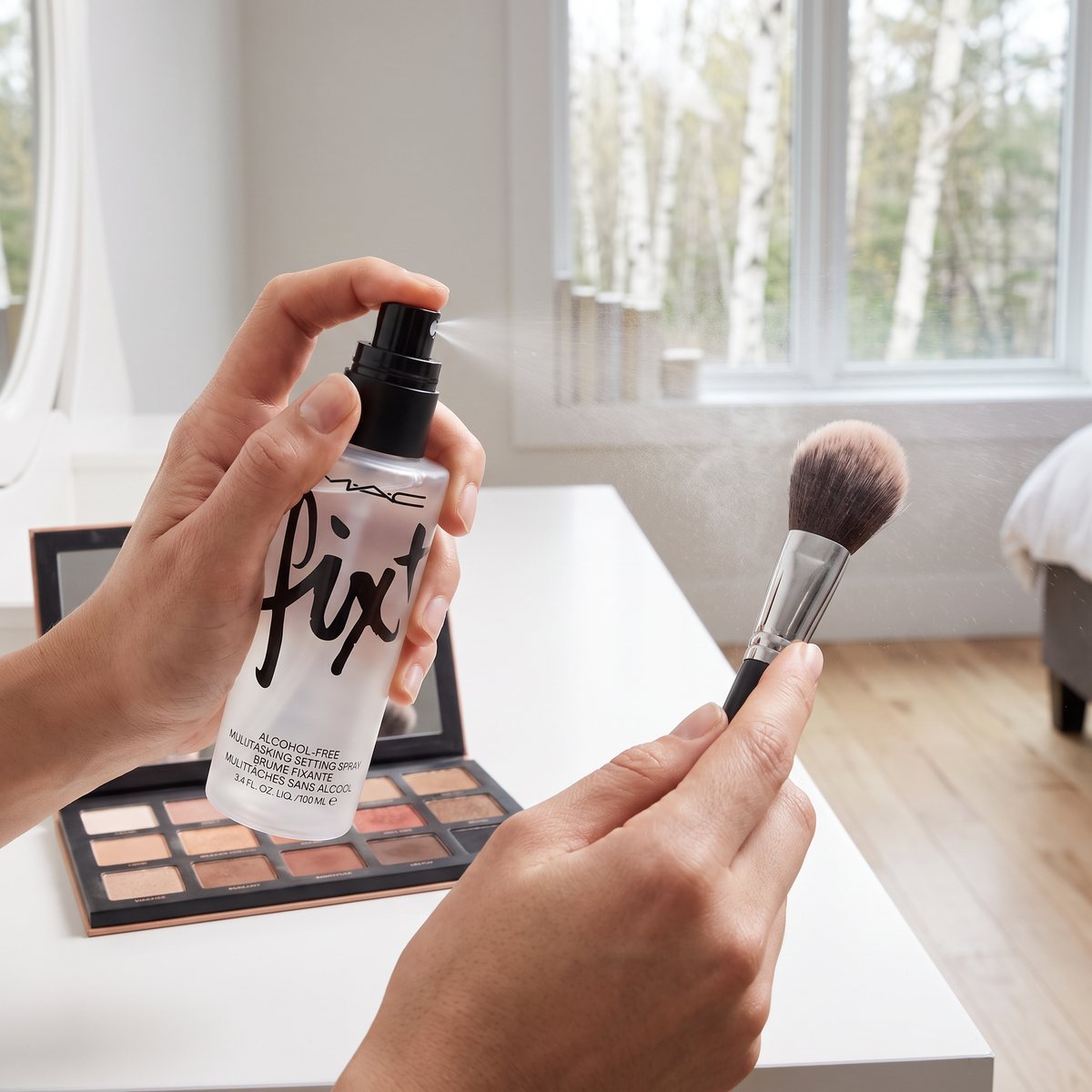 MAC Fix+ sprayas på en ögonskuggspensel ovanför en öppen ögonskuggspalett vid ett vitt sminkbord