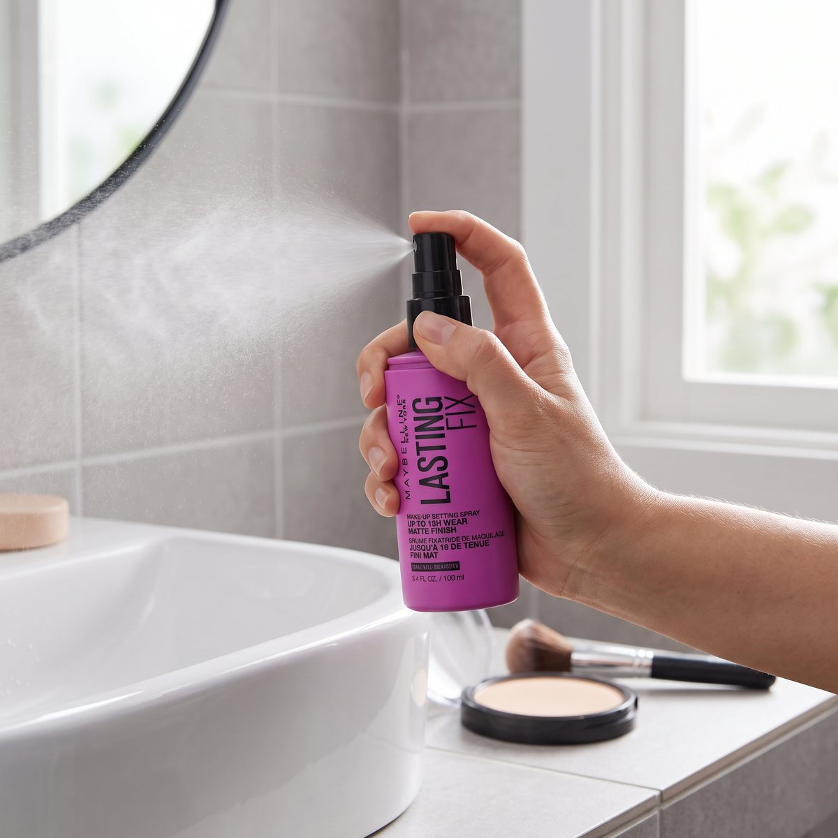 Hand som sprayar Maybelline Lasting Fix vid ett vitt handfat med puderdosa i ett ljust badrum