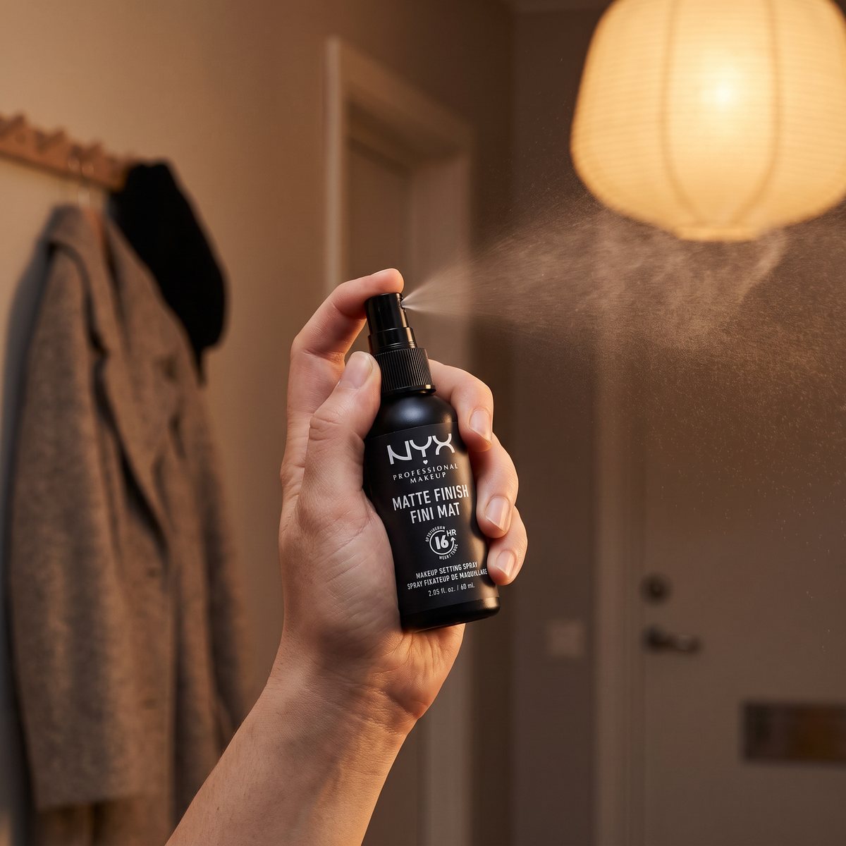 Hand som sprayar den lilla svarta NYX Matte Finish-flaskan i en hall med kappa på kroken