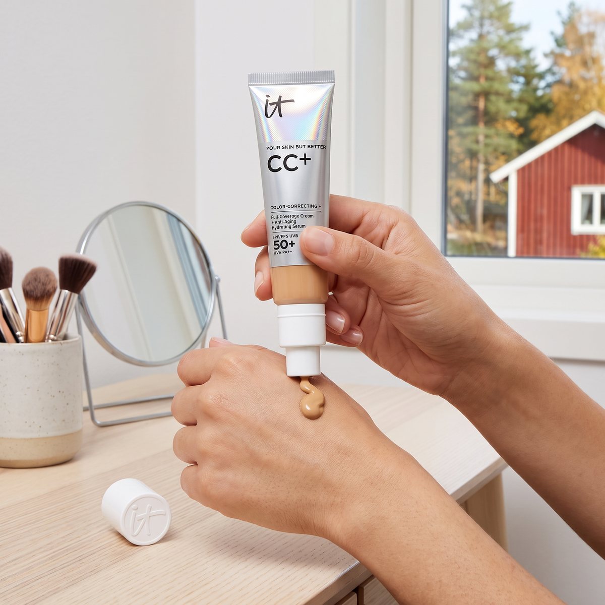 IT Cosmetics CC+ Cream kläms ut på handryggen vid ett sminkbord av björk, med tallar och en röd stuga utanför fönstret