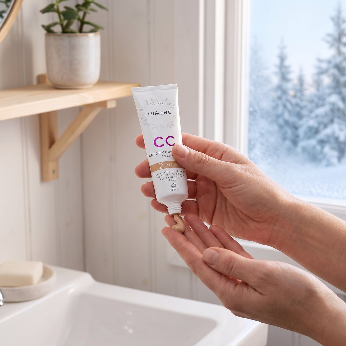 Lumene CC Color Correcting Cream kläms ut på fingrarna i ett svenskt badrum med vit panel och snötäckt granskog utanför fönstret
