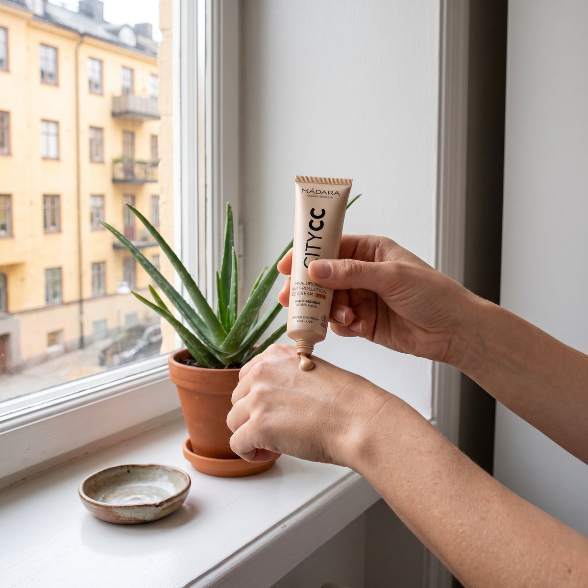 Mádara City CC klicks ut på handryggen vid ett fönster med aloe vera i kruka och gula stenhusfasader utanför