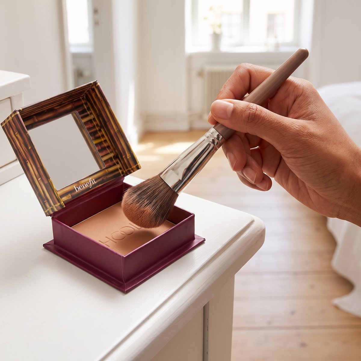 Borste doppas i Benefit Hoola Matte Bronzer som står öppen på en vit sminkbyrå i ett ljust svenskt sovrum