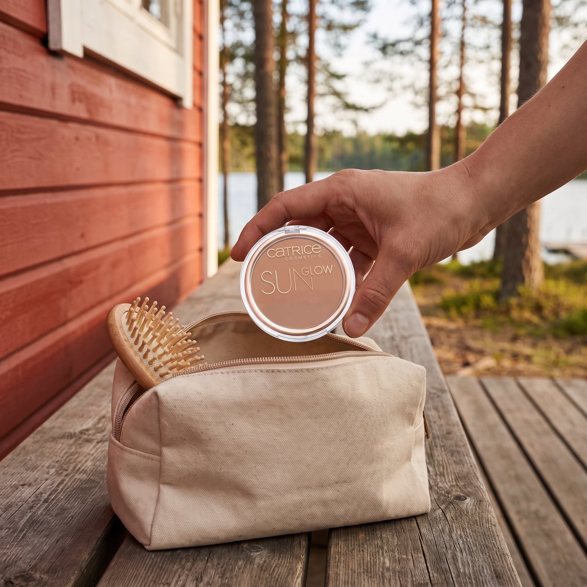 Hand lyfter Catrice Sun Glow Matt Bronzing Powder ur en necessär på en träbänk vid en röd stuga vid en sjö