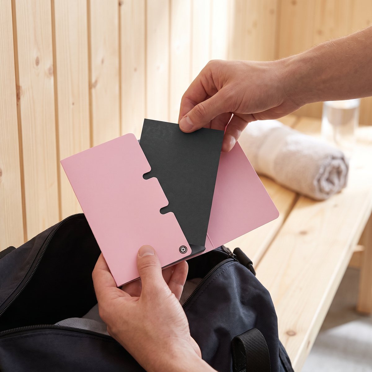 Händer tar ett svart kolark ur Brushworks rosa häfte ovanför en träningsväska i ett omklädningsrum med ljus träpanel