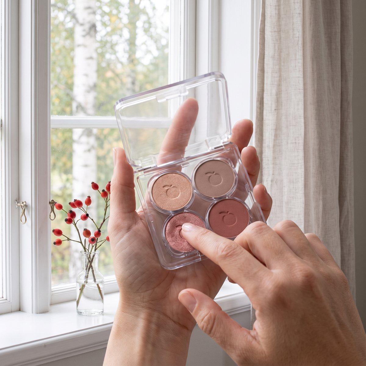 IDUN Minerals Eyeshadow Palette Nyponros öppen i handflatan medan ett finger tar upp den roséguldskimrande nyansen vid ett vitt spröjsfönster med björkar utanför