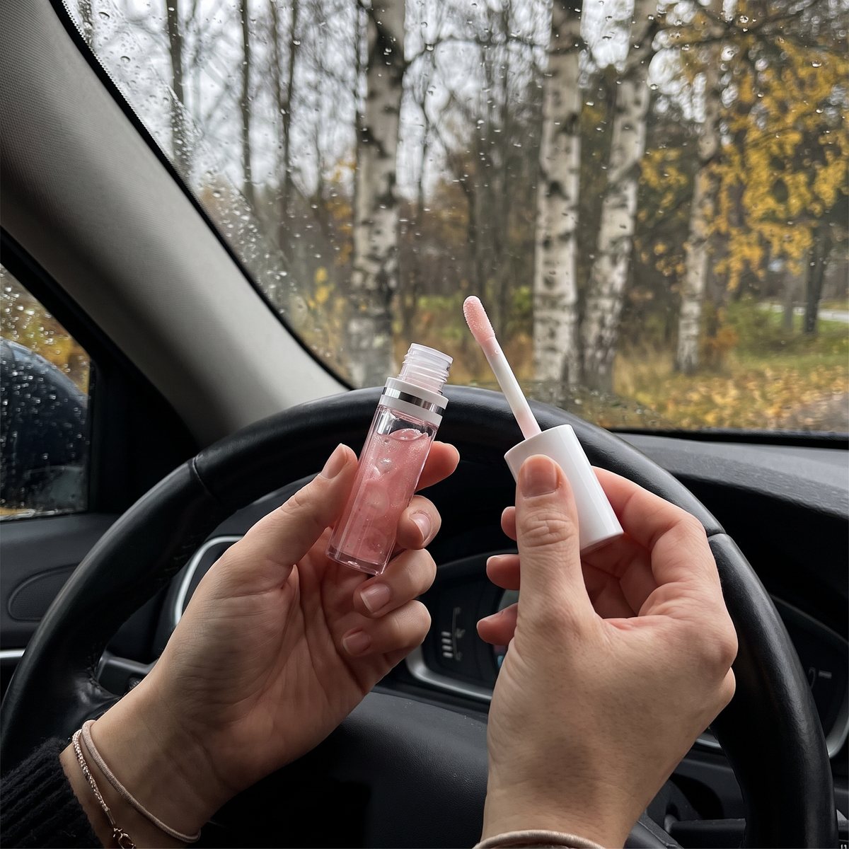 Händer håller Labello Lip Oil Dress Nude med skimrande rosa olja och applikatorn i en parkerad bil en regnig höstdag med björkar utanför