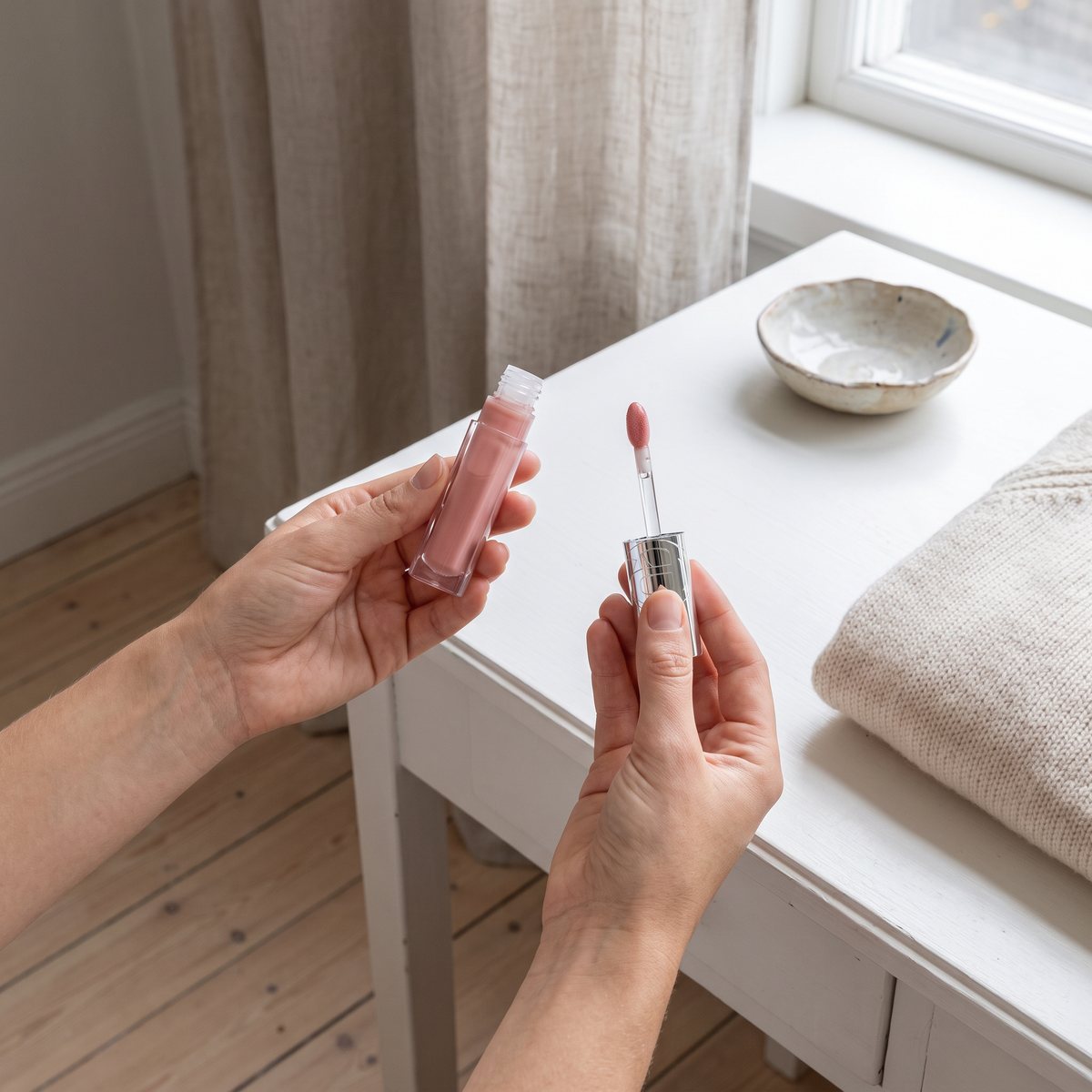 Händer håller L'Oréal Paris Plump Ambition Hyaluron Lip Oil 650 Nude Macaron och den rundade applikatorn vid ett vitt sminkbord i ett svenskt sovrum