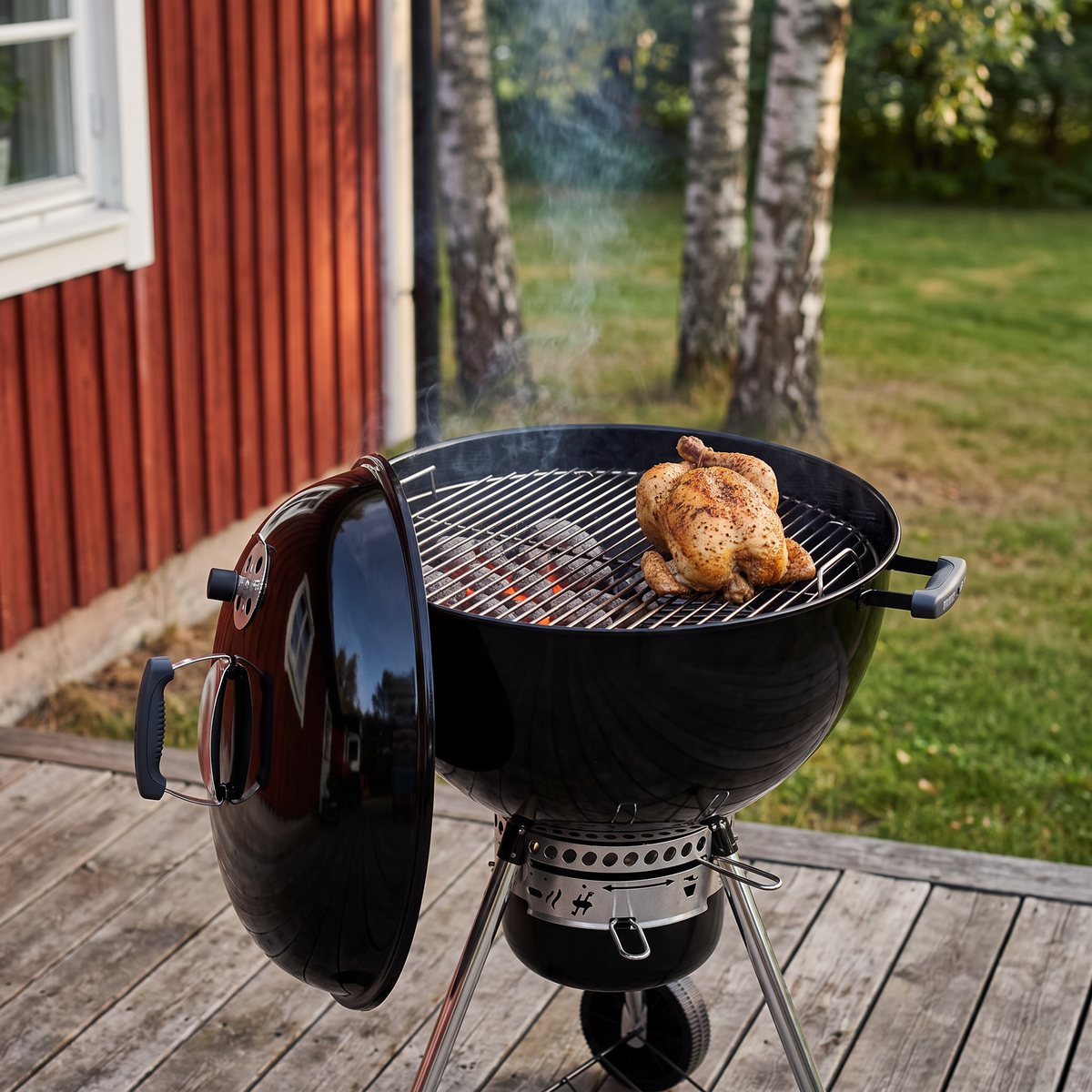 Weber Master-Touch klotgrill med locket upphängt på sidan och en hel kyckling på den svala halvan av gallret, på en trädäck vid ett rött trähus med björkar