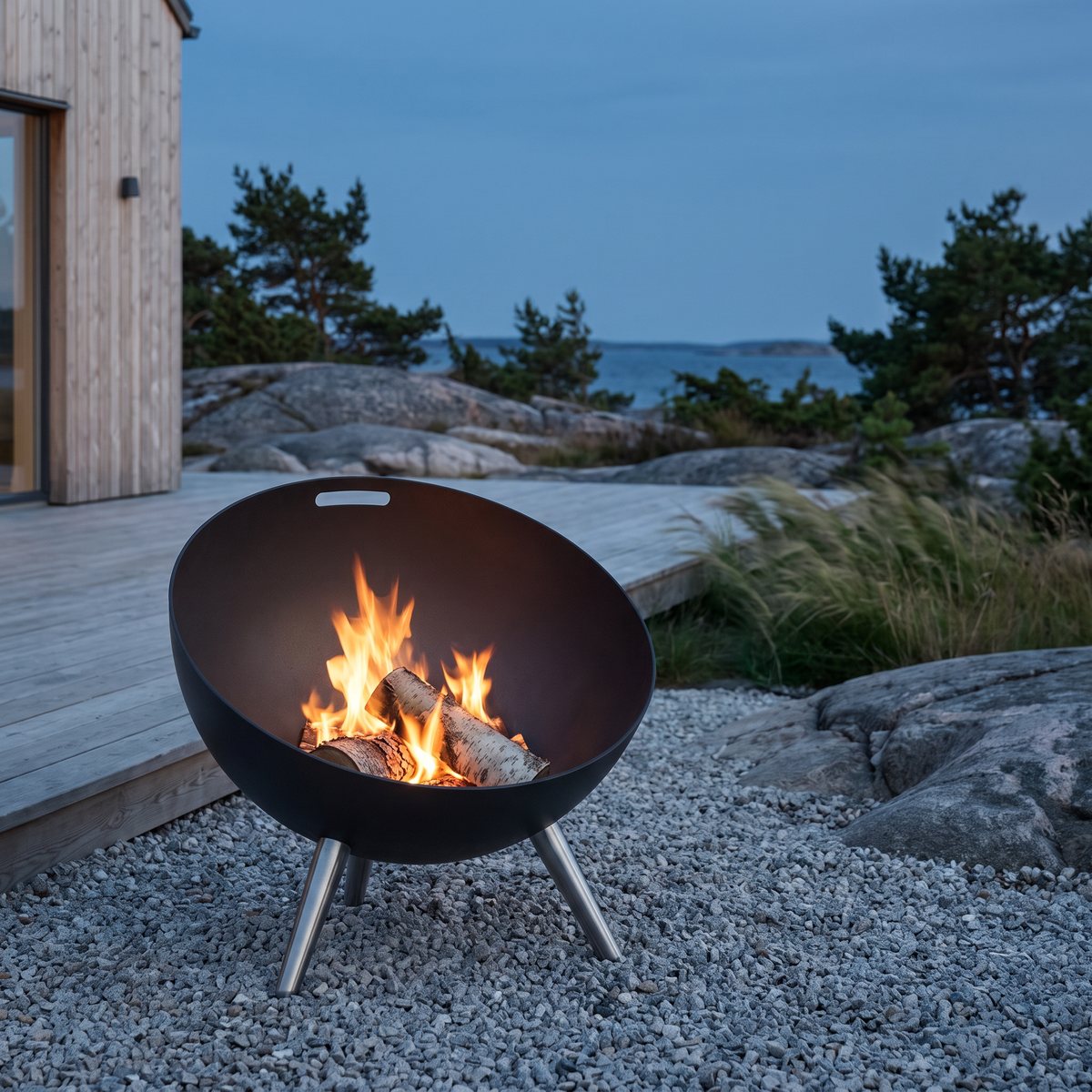Eva Solo FireGlobe med brinnande björkved på grus vid ett trähus i skärgården i skymningen