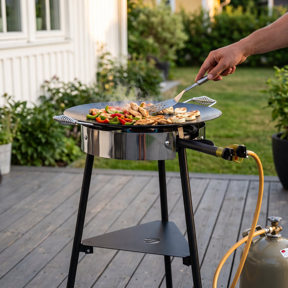 Hällmark HM8701 V2 stekhällsset med gasolbrännare på stativ på en trädäcksaltan, med kyckling och paprika på hällen