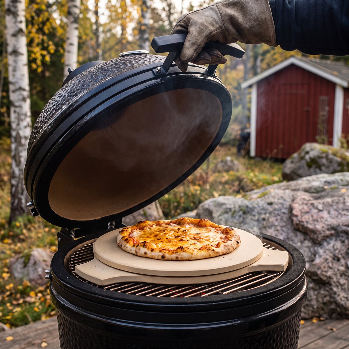 Beige pizzasten från Kamado Sumo med pizza i en öppen kamadogrill på en svensk tomt med björkar och en röd bod