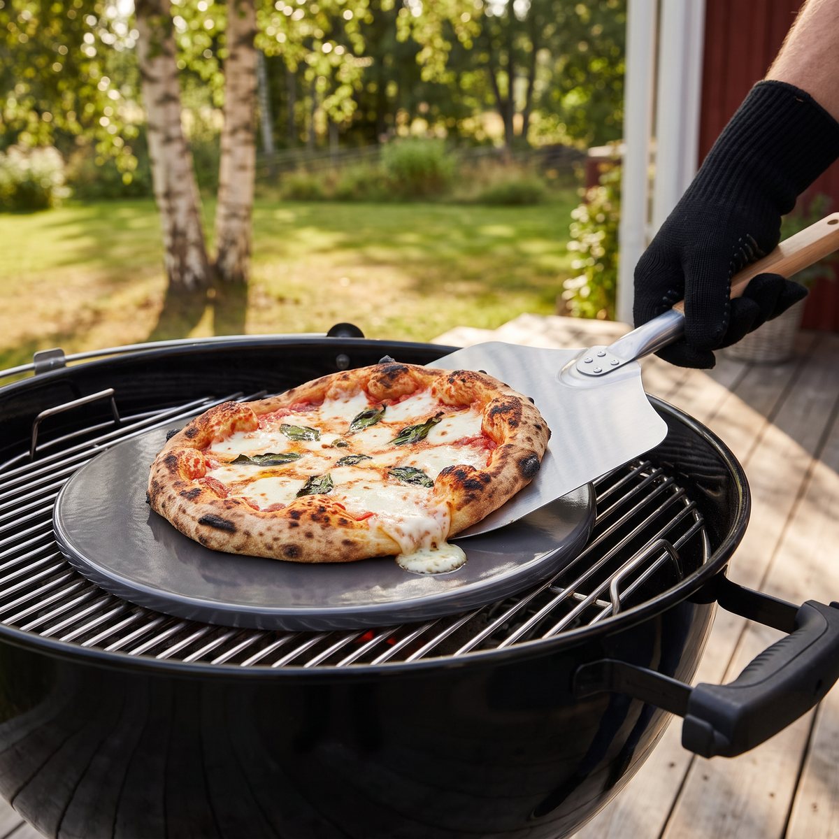 Mörkgrå glaserad pizzasten från Weber med pizza på gallret i en klotgrill på en altan i en svensk trädgård med björkar