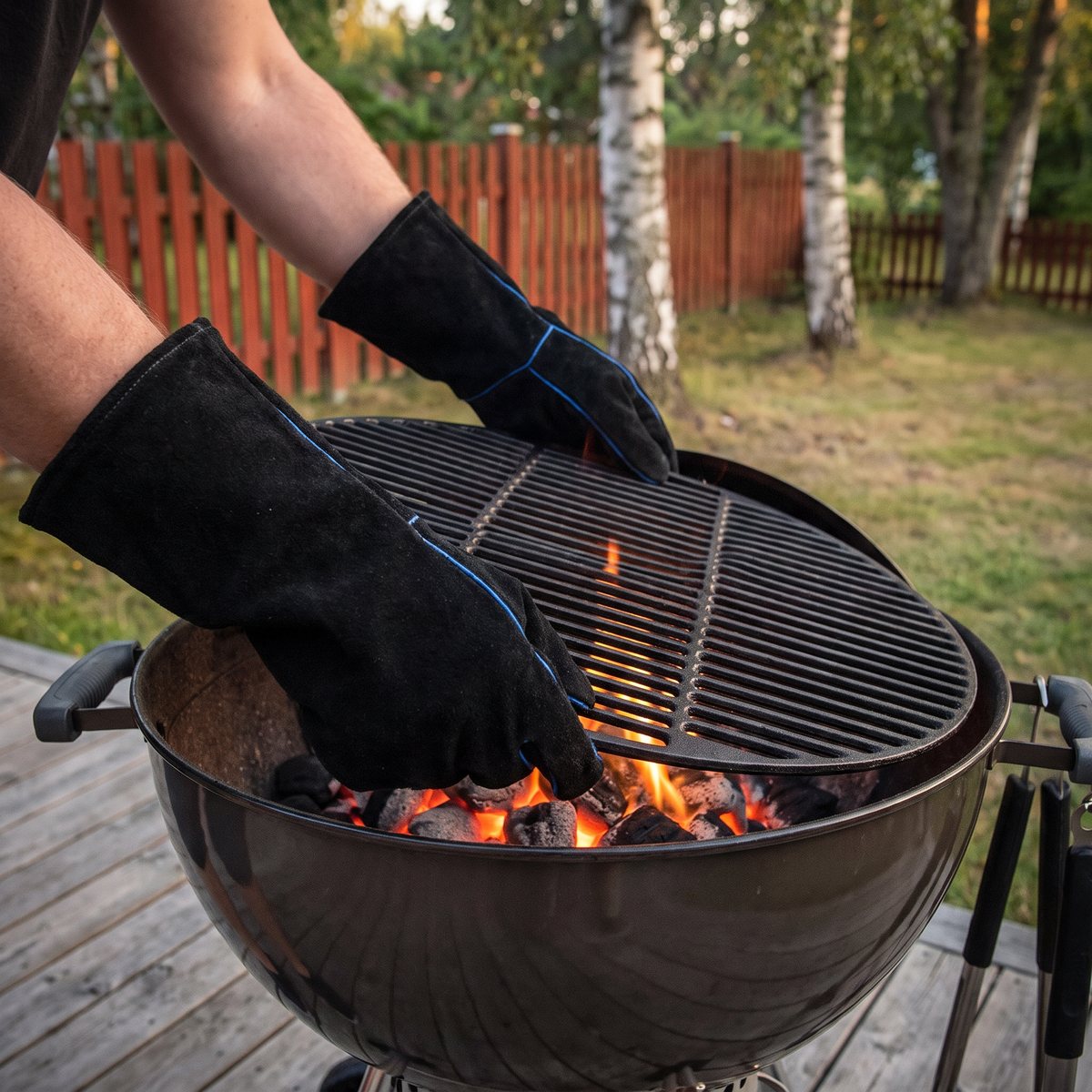 Svarta Broil King-grillhandskar i läder med långa manschetter håller i gjutjärnsgallret på en klotgrill med glödande kol, på en trädäck i en svensk trädgård med björkar och rött staket