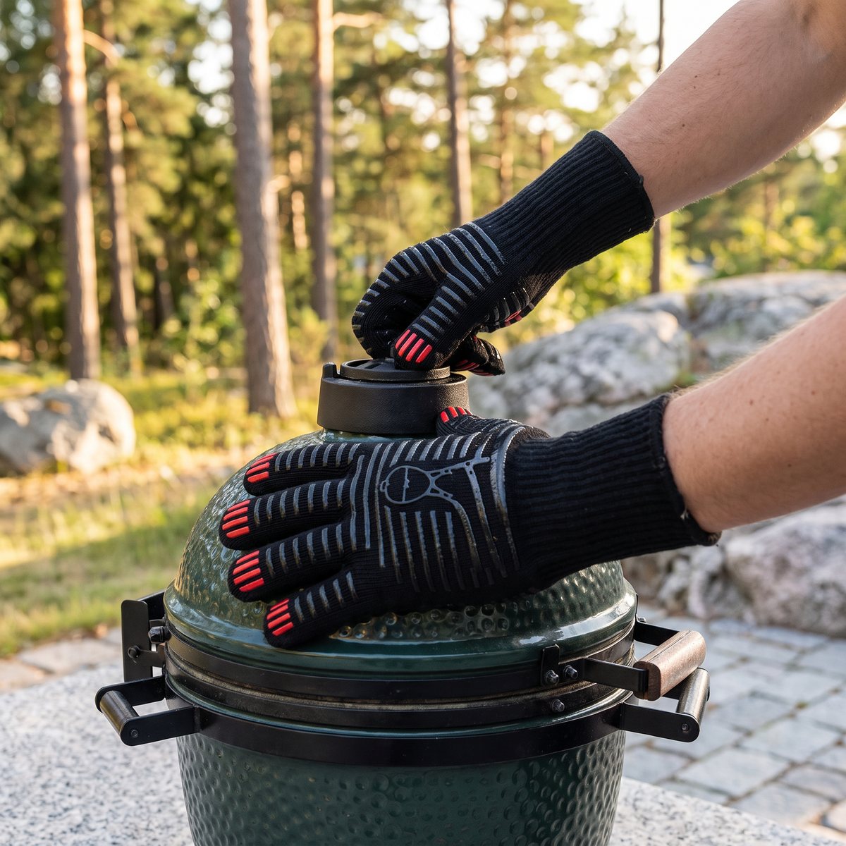 Svarta Kamado Sumo-grillhandskar i tyg med röda och grå silikongrepp vrider på spjället på en grön kamadogrill, på en stenlagd uteplats bland tallar och granithällar