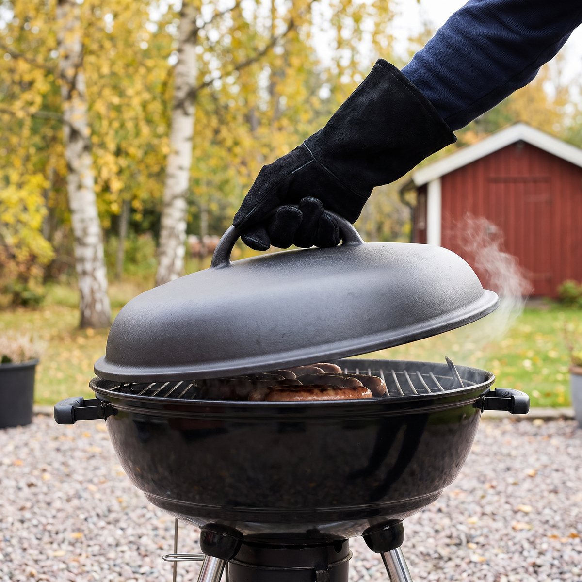 Svart Morsø-grillhandske i mocka på höger hand lyfter locket på en kolgrill med korv, på en grusgång i en svensk trädgård om hösten