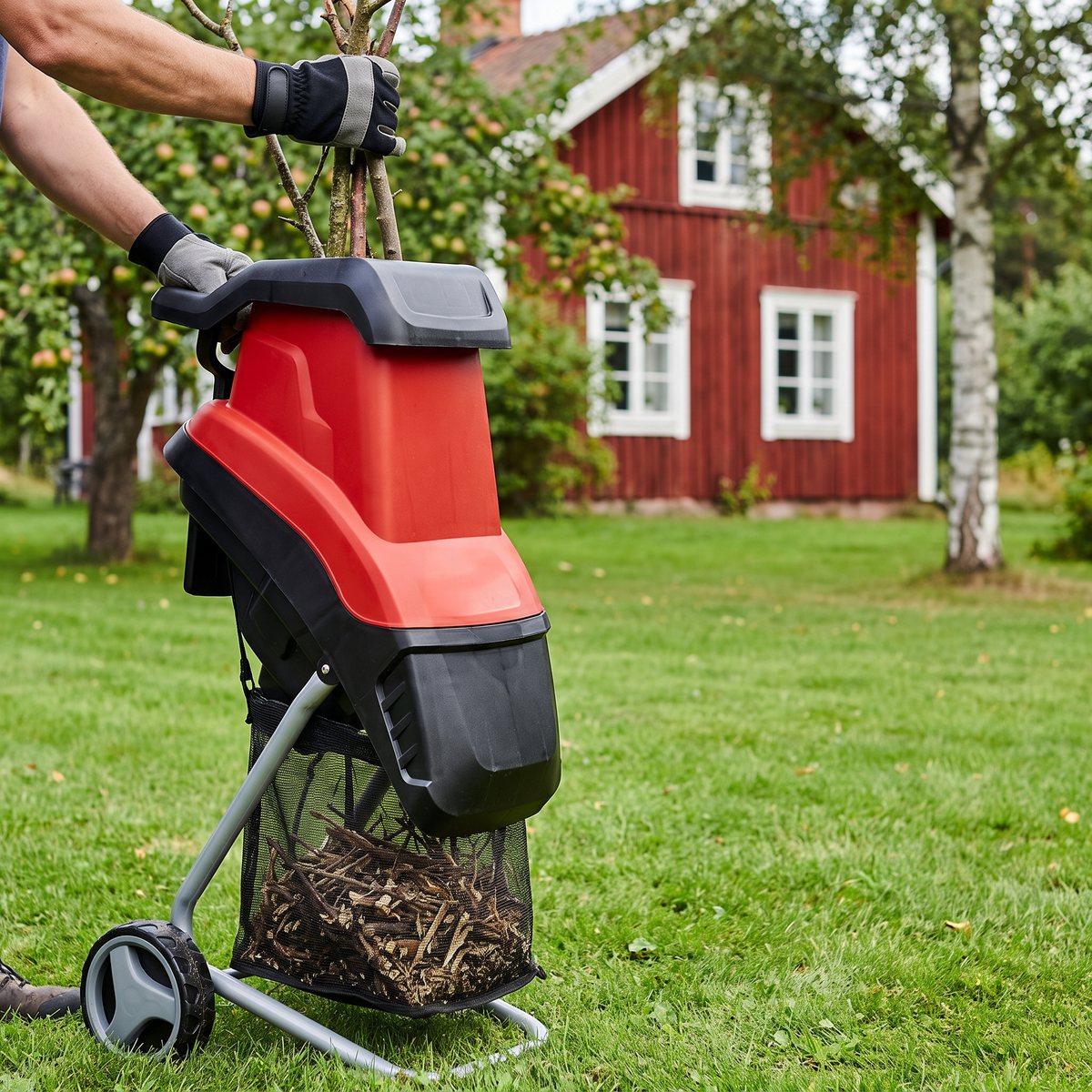 Einhell GC-KS 2540, röd och svart kompostkvarn med uppsamlingspåse, på gräsmattan vid en röd stuga med fruktträd och björk