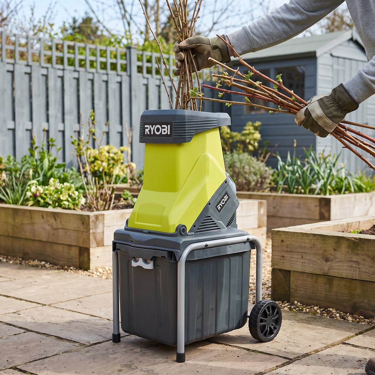 Ryobi RSH2545B, limegrön kompostkvarn med grå uppsamlingslåda, på en stenlagd uteplats bland odlingslådor där hallonris matas ner i tratten