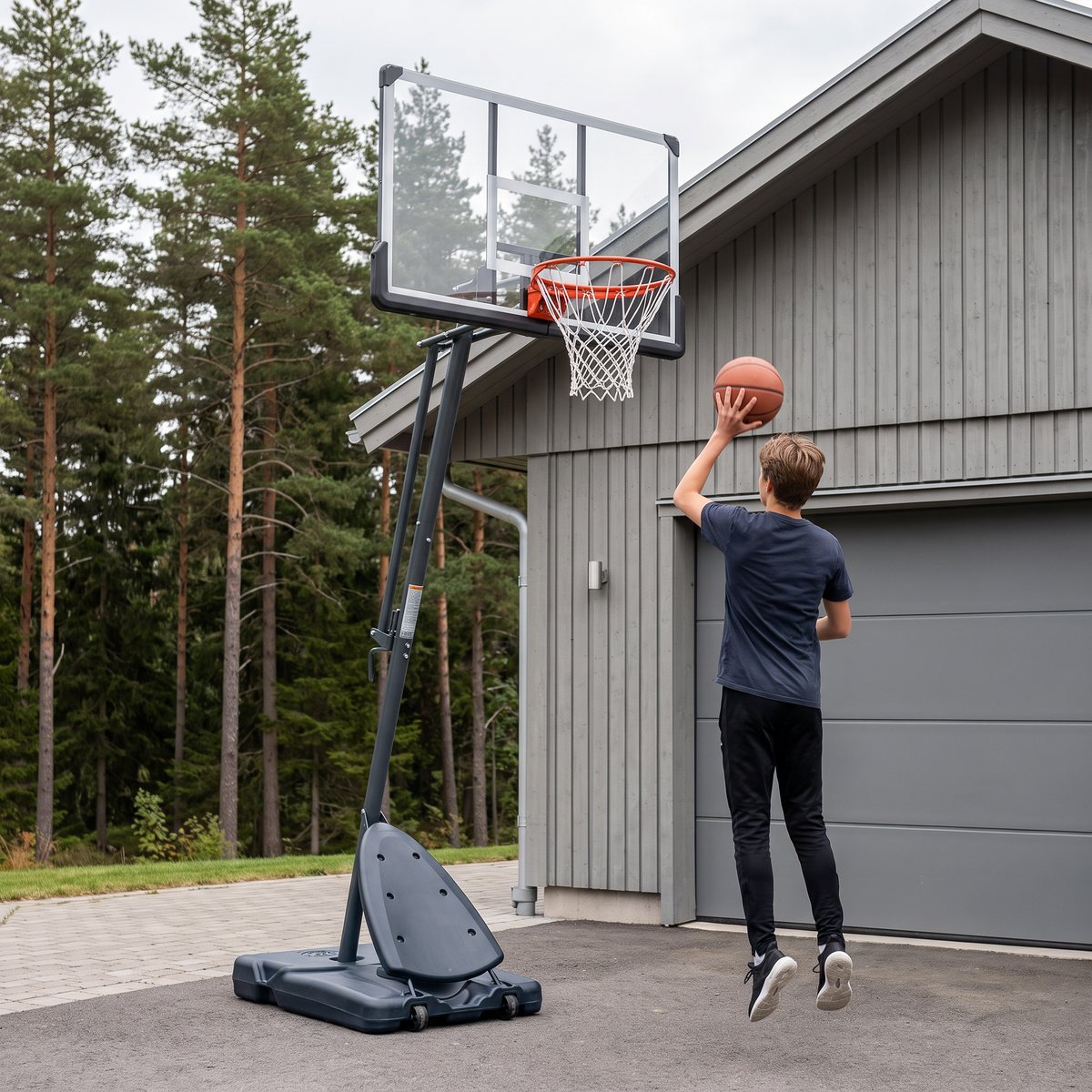 FitNord Hoop 500 på en asfalterad uppfart vid ett grått trähus med garage och tallskog, där en tonåring hoppar upp mot korgen med bollen.