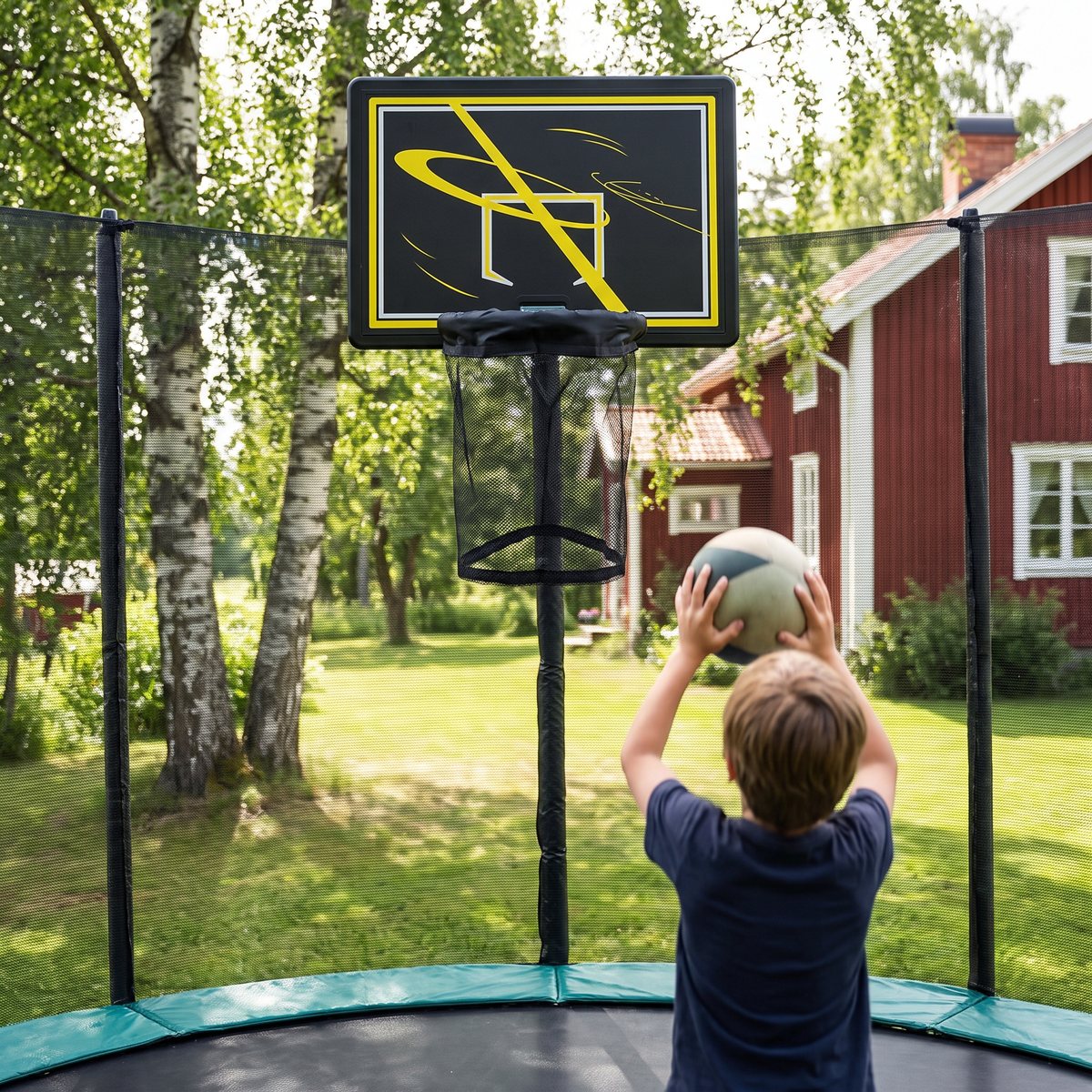 ProSport Basketkorg för studsmatta fäst på skyddsnätets stolpe, med ett barn som kastar en mjuk boll från studsmattan i en svensk trädgård.