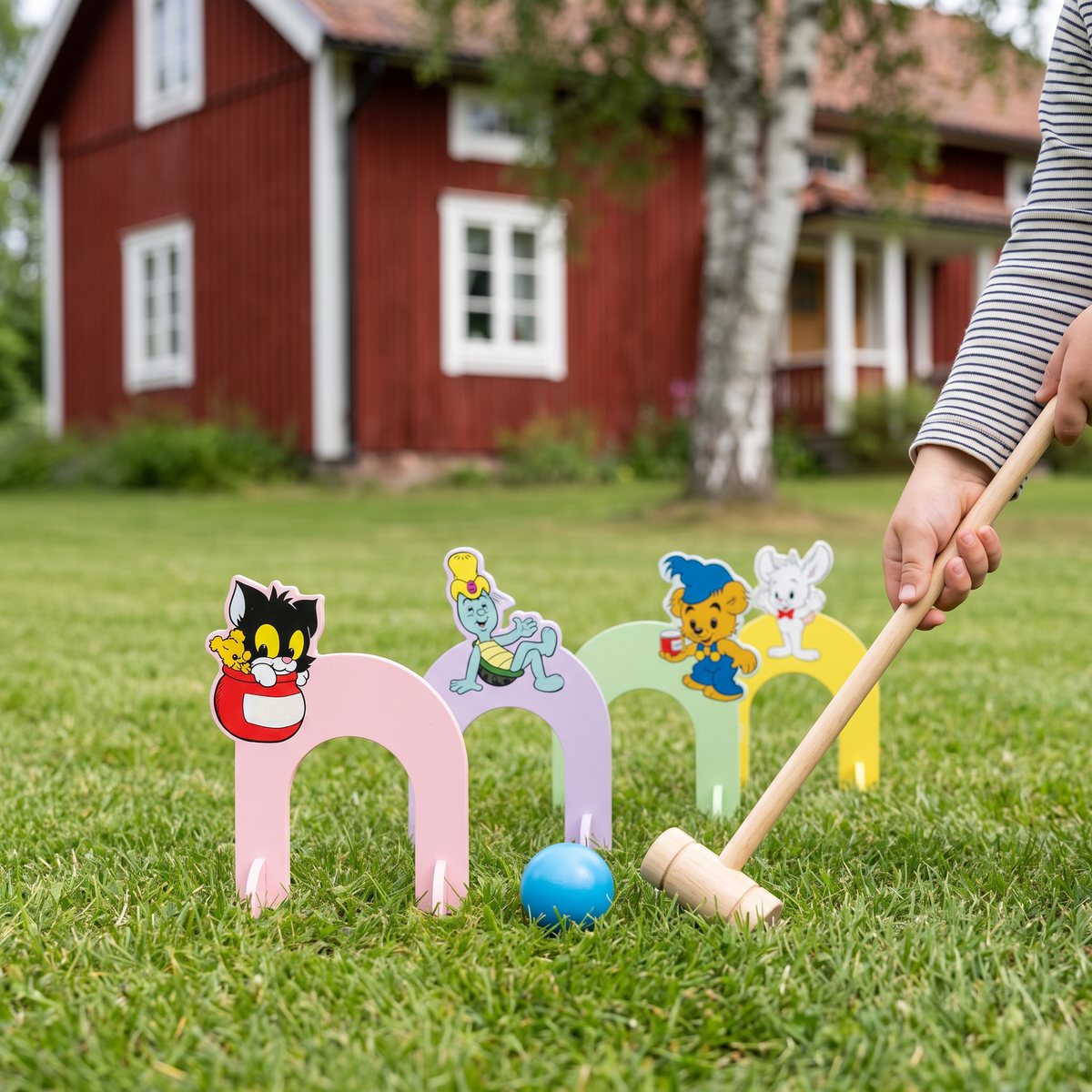 Bamse Krocketspel med pastellfärgade bågar och seriefigurer uppställda på en gräsmatta framför ett rött svenskt trähus, där ett barn slår den blå bollen med en kort träklubba