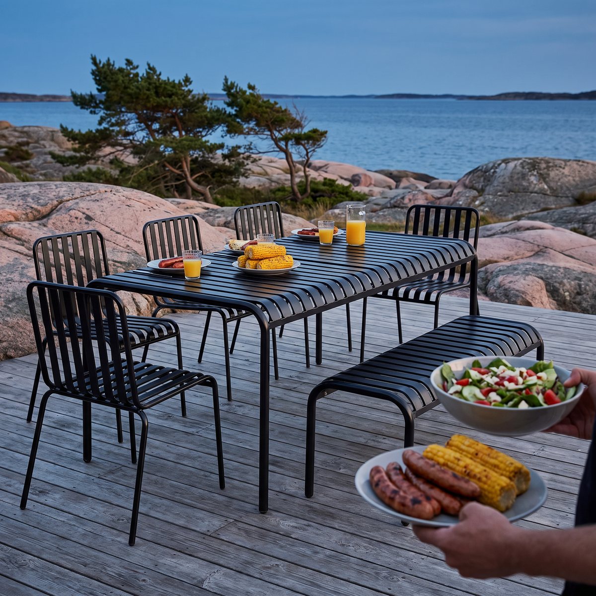 Lyfco Bergvik, svart matgrupp i stål med långbord, fyra stolar och bänk, dukad för grillkväll på ett trädäck vid västkustens klippor.
