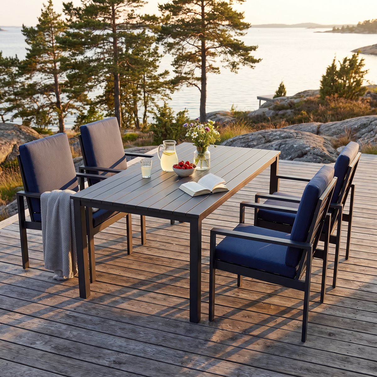 Lyfco Hamnviken, svart matgrupp i aluminium med fyra stolar och djupblå dynor, på ett trädäck vid skärgården i kvällssol.
