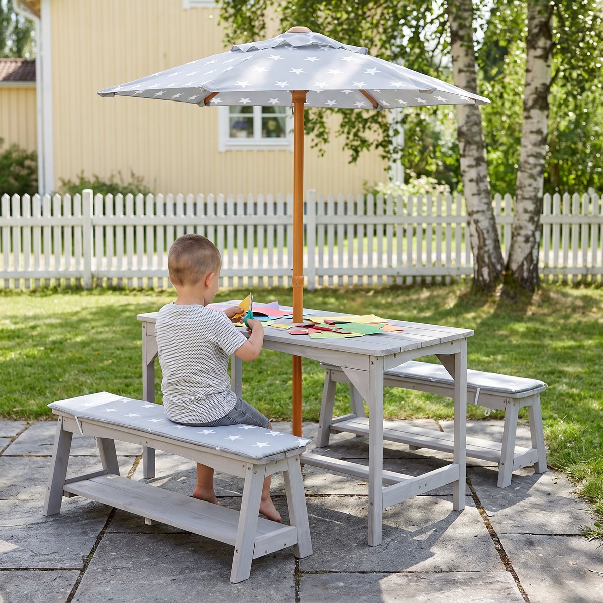 Grått Roba picknickset med parasoll och stjärnmönstrade dynor på en stenlagd uteplats, där ett barn klipper färgat papper