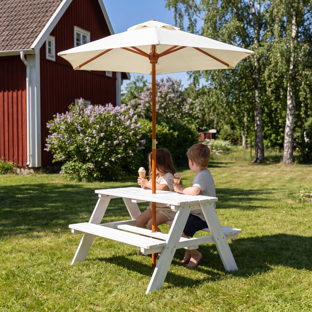 Vitt Woodlii picknickbord med uppfällt parasoll i en svensk trädgård med syren och rött trähus, där två barn äter glass i skuggan
