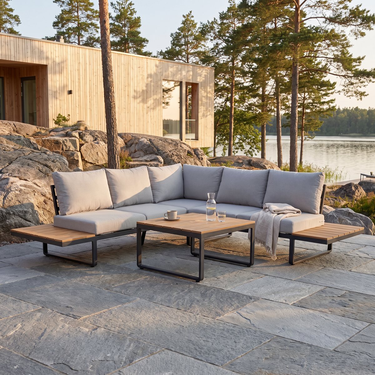 Låg hörnsoffa i svart aluminium med ljusgrå dynor och bord med polywoodskiva på en stenlagd terrass vid ett nybyggt svenskt hus, tallar och sjö i kvällssol