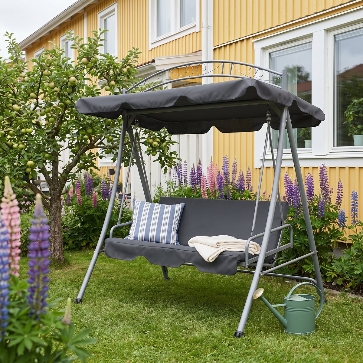 Antracitgrå vidaXL-hammock med dekorativ båge och en sammanhängande dyna på gräsmattan framför ett gult svenskt radhus med lupiner