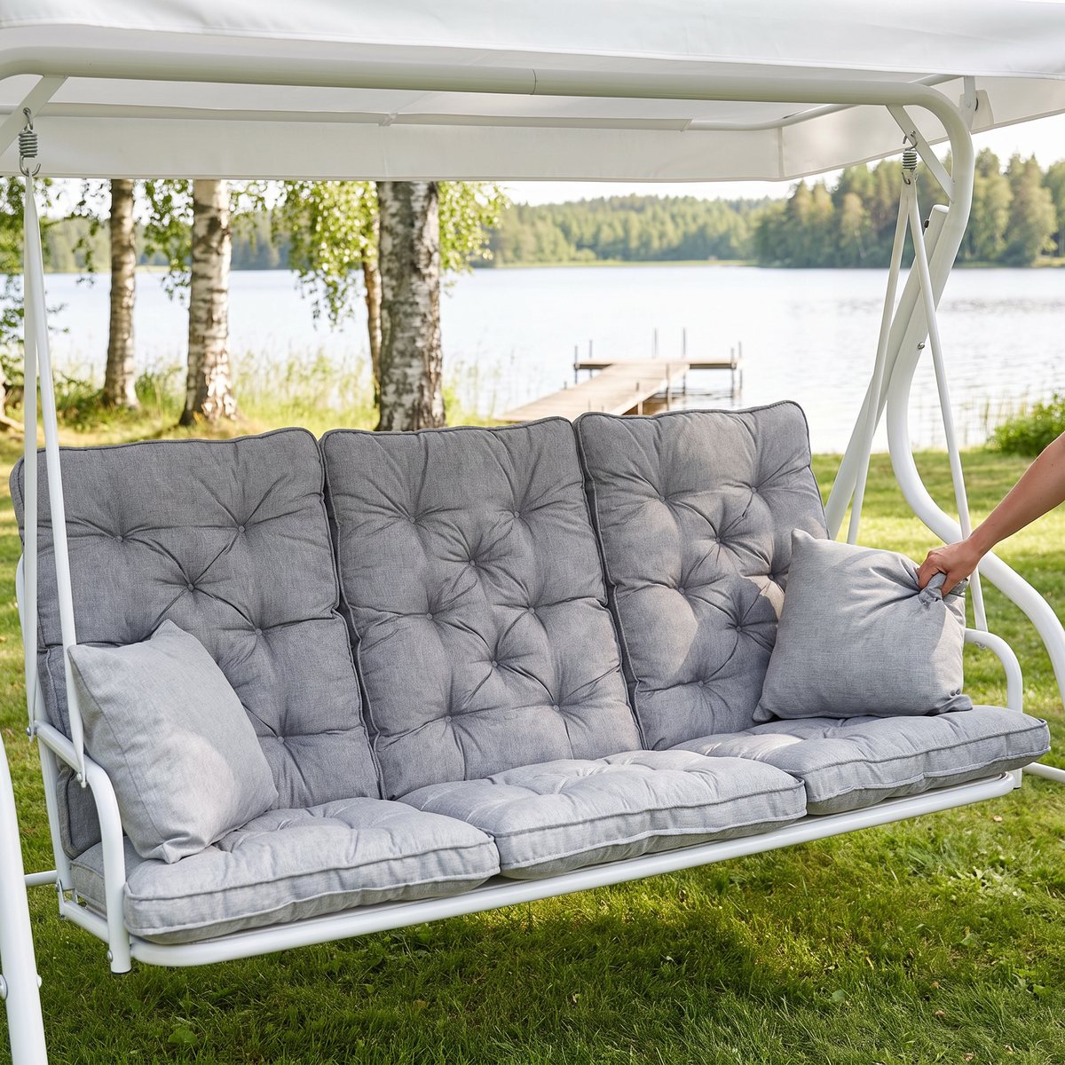 Grått dynset med tre knappade ryggdynor och två sidokuddar i en vit hammock på en gräsmatta vid en sjö med brygga och björkar