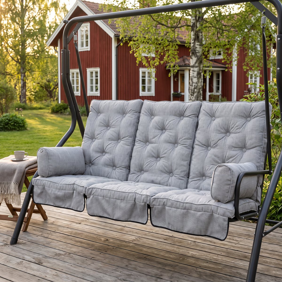 Ljusgrå boxsydda hammockdynor med hög rygg och två sidokuddar i en hammock på en trätrall framför ett falurött hus med björkar