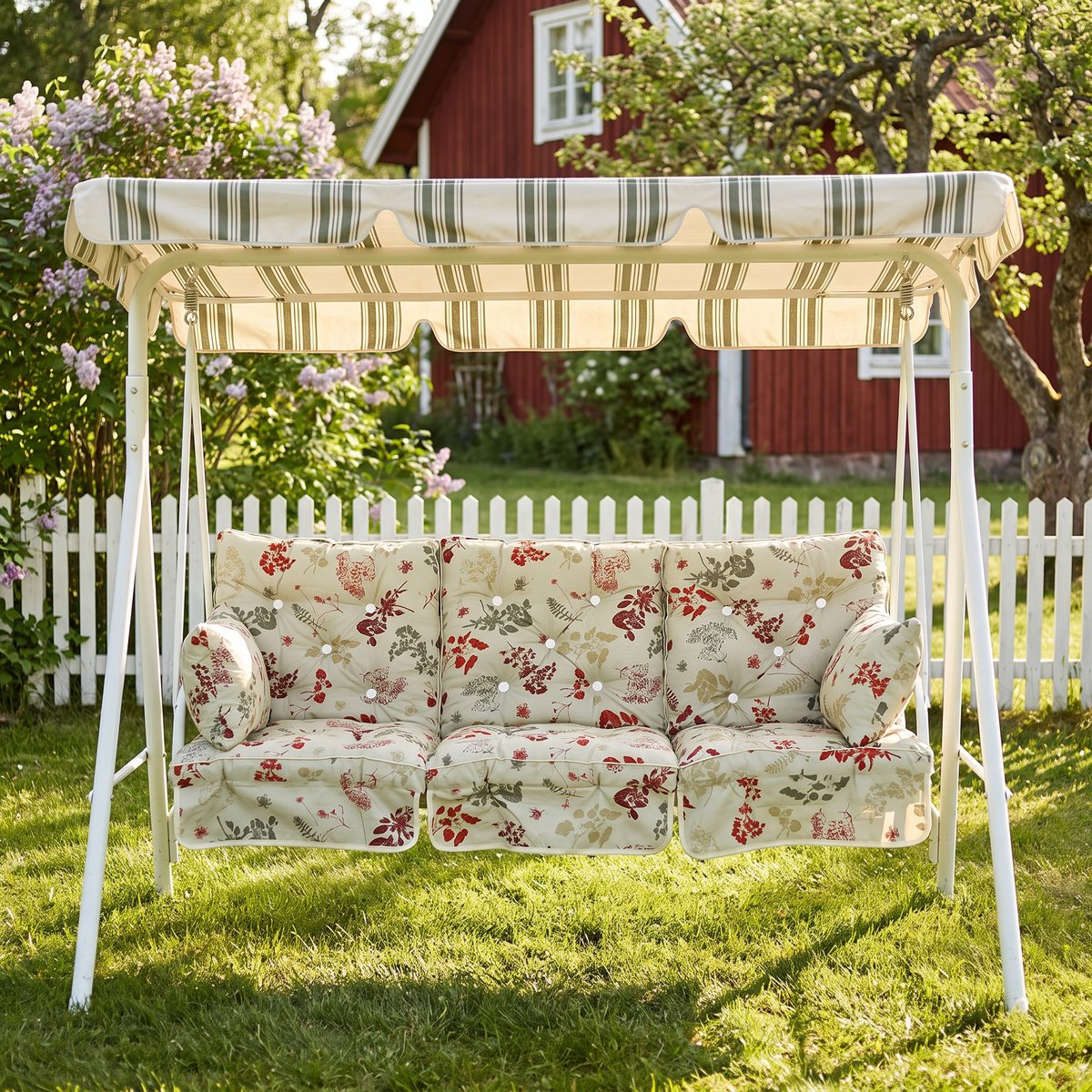 Blommönstrat dynset med låg rygg och sidokuddar i en vit hammock med randigt tak i en trädgård med syren, vitt staket och rött torp