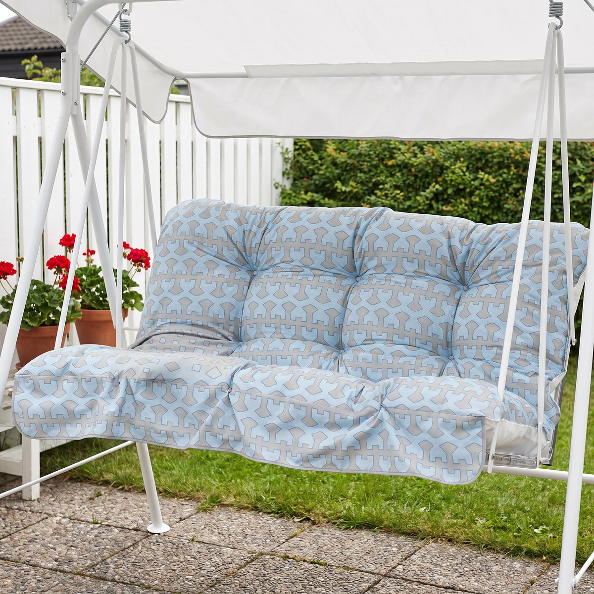Ljusblått mönstrat dynset i en tvåsitsig vit hammock med tak på en stenlagd uteplats med pelargoner och vitt staket