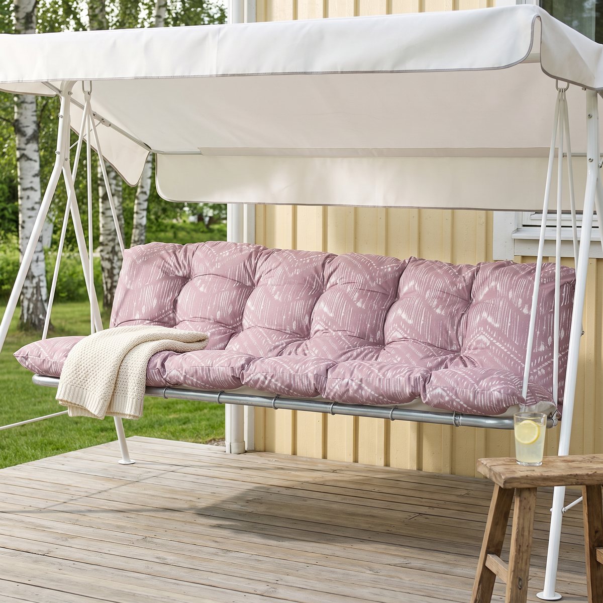 Rosa dynset med sittdyna och ryggdyna i en vit hammock med tak på en trätrall vid ett gult trähus med björkar