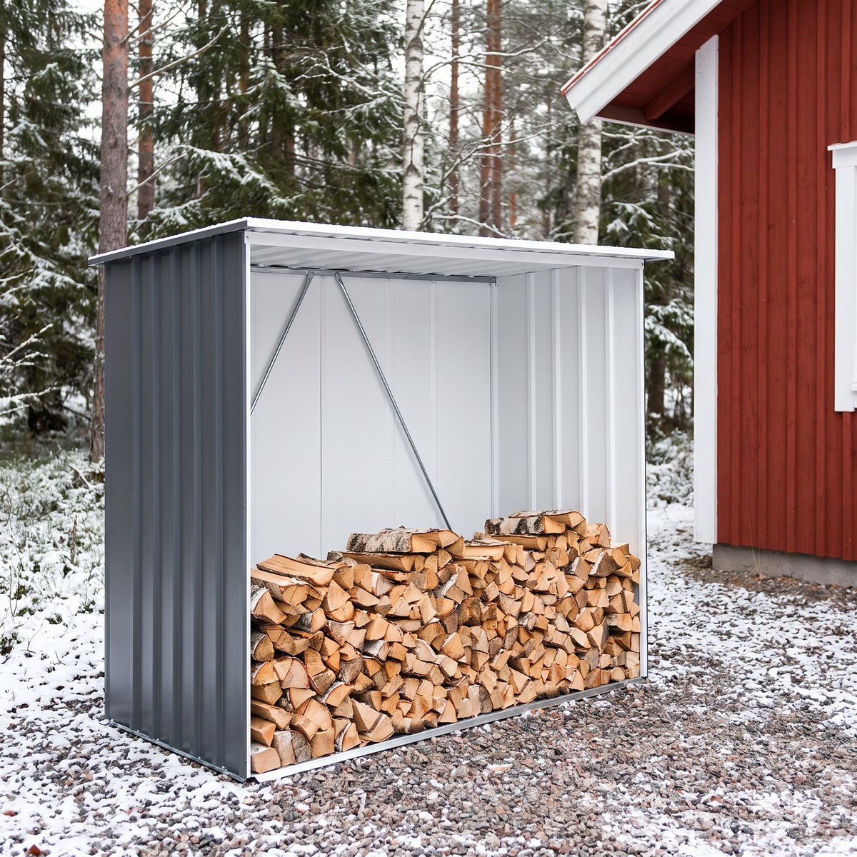 Mörkgrått vedskjul i plåt med vit insida, fyllt med björkved, på grusplan vid ett rött trähus i snöig skog