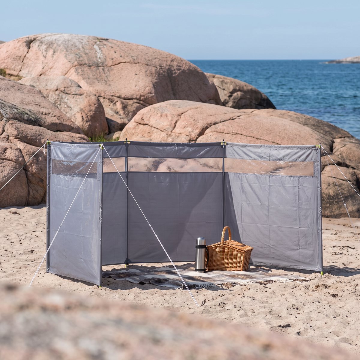 Grått vindskydd uppspänt med stolpar och linor på en sandstrand vid granitklippor och hav, med filt och picknickkorg i lä