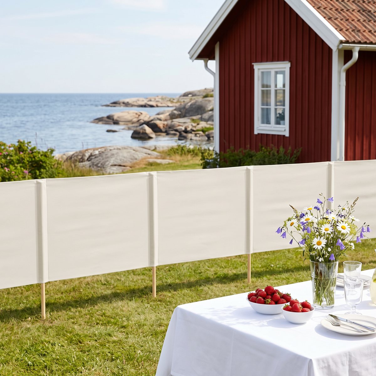 Sandvitt vindskydd med fem tygpaneler mellan trästolpar på en gräsmatta vid en röd stuga i skärgården, med dukat bord