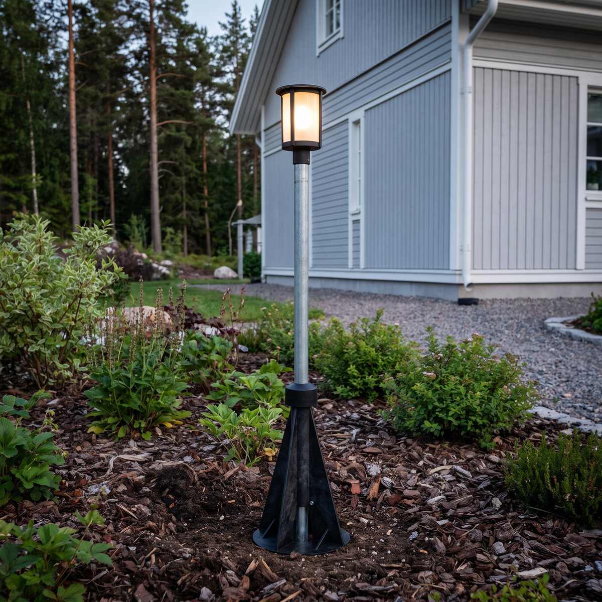 Garo MVRF stolpfundament med rörstolpe och en trädgårdslykta överst, i en rabatt framför ett grått trähus