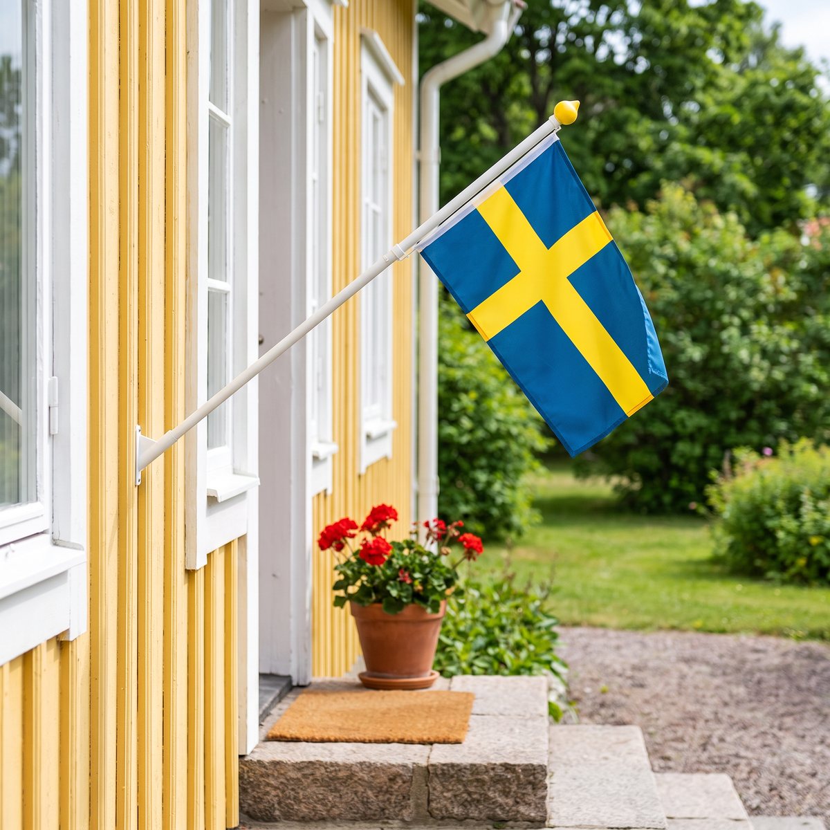 Liten vit fasadflaggstång med svensk flagga vid entrétrappan på ett gult trähus med pelargoner