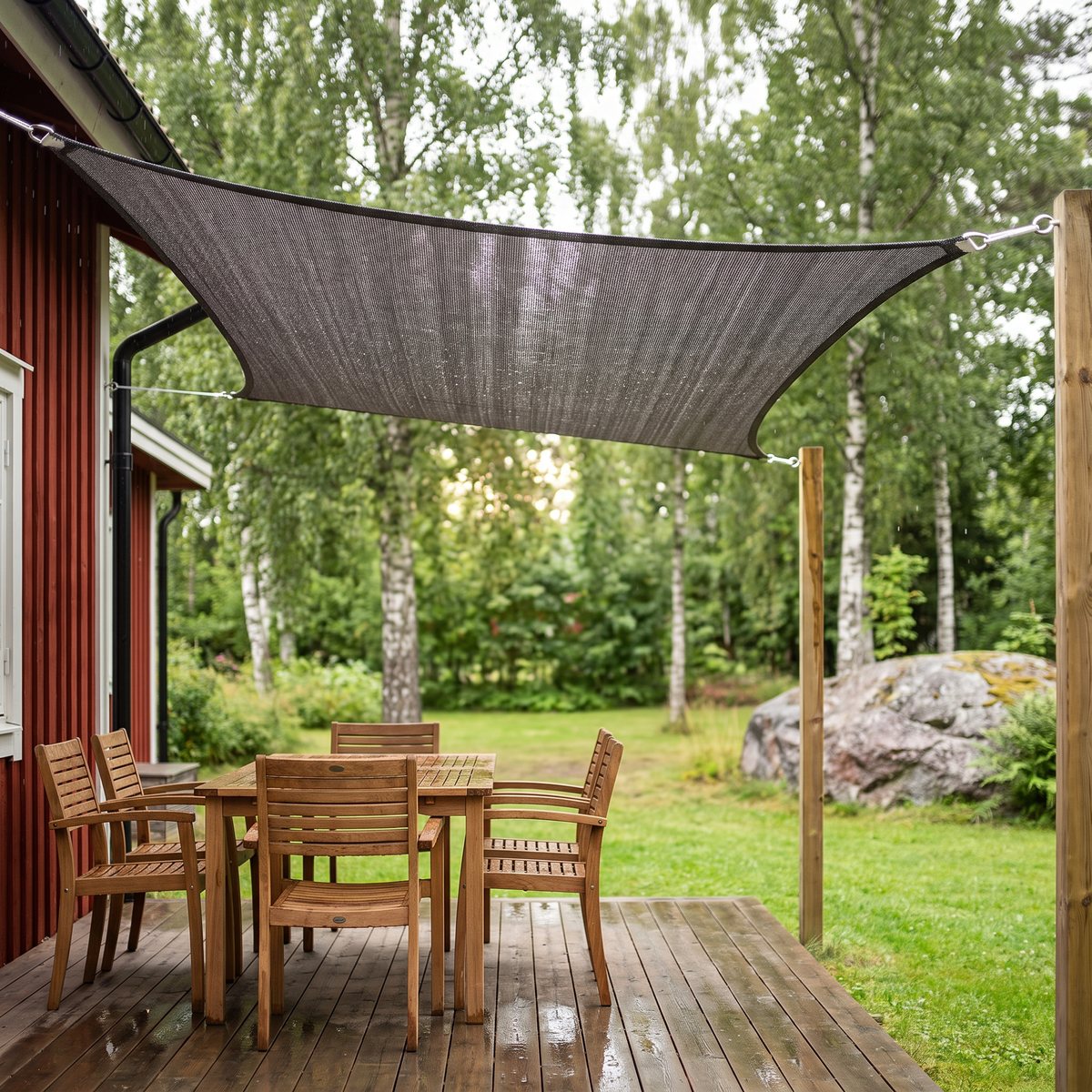 Antracitgrått fyrkantigt solsegel i HDPE spänt mellan ett falurött trähus och två trästolpar över en blöt trätrall med matbord, svensk trädgård med björkar